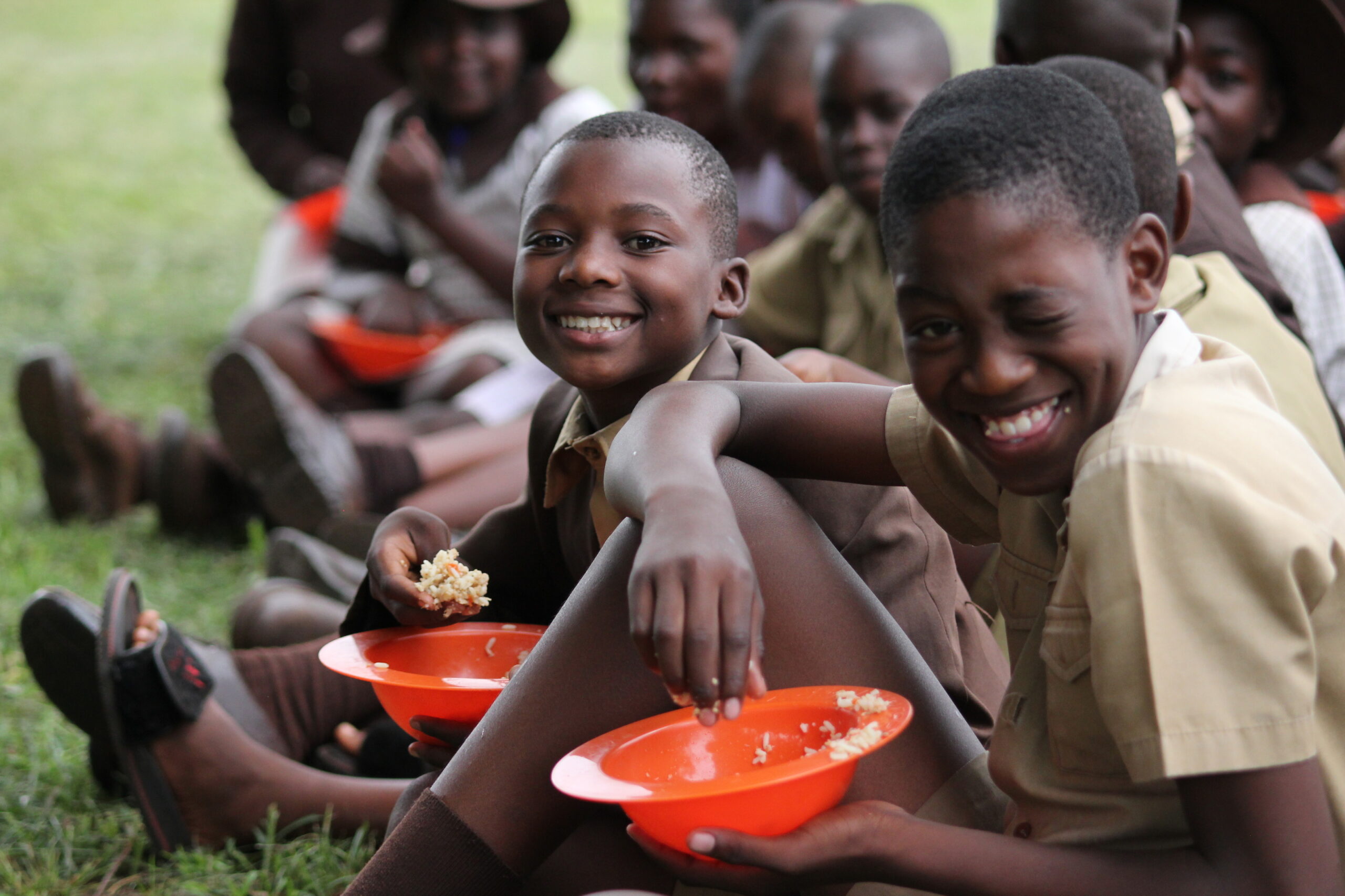 Some Zimbabwean boys in school uniform are sitting on green grass eating their lunch out of orange bowls. The boy in the foreground winks to the camera; the child next to him smiles broadly at the camera.