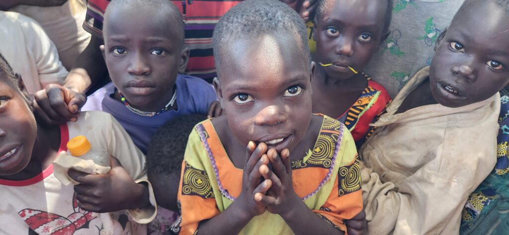 A young child from Uganda, surrounded by their friends, smiles up a the camera. They are wearing an orange top and their hands are clasped together by their mouth.