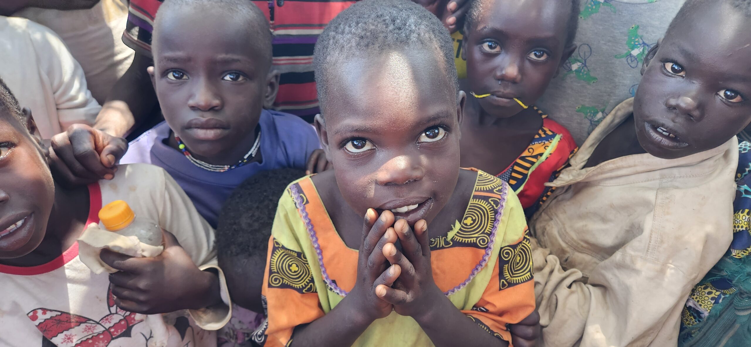 Some Ugandan children look straight up into the camera. The child in the middle is wearing an orange and yellow Ankara print top. She has her hands up by her mouth and is looking straight up with large brown eyes. The children all look dishevelled.