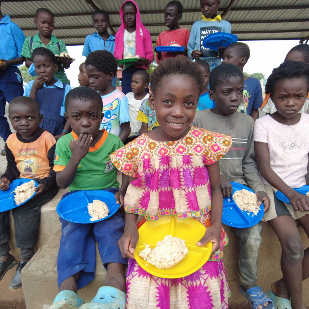 A young Zambian girl in a bright pink and yellow dress smiles at the camera. Three rows of children are behind her, and they're all holding plates loaded with nutritious food.