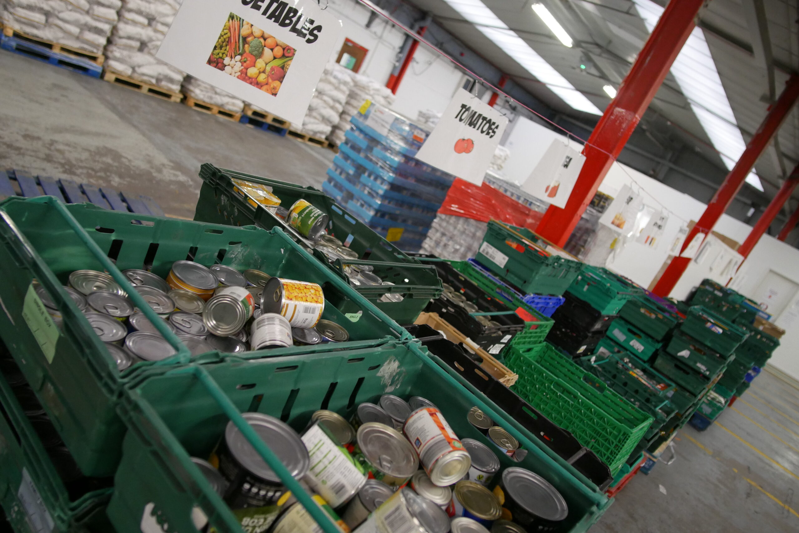 A warehouse stacked with green and blue delivery trays, some of which are full. Signs above the trays say things like 'vegetables' and 'tomatoes'. The food is sorted into the trays below the signs.