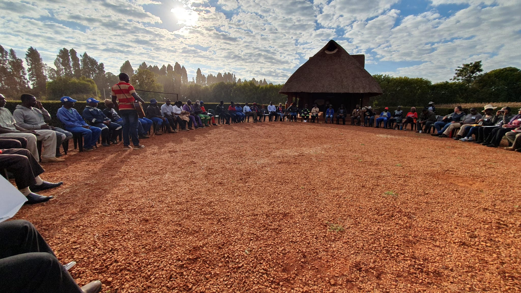 A community of farmers gather in a circle to learn sustainable farming techniques. They're sat outside on green plastic chairs. In the background is a traditional thatched hut.