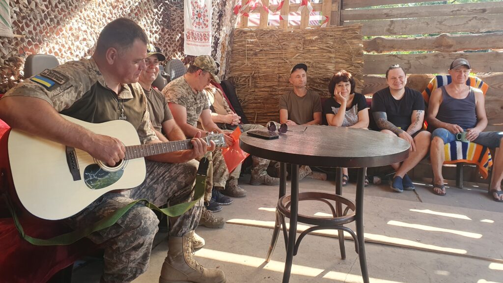 A man in combat gear plays the guitar in a small basic hut. Sitting in a semi circle beside him are a group of other men in combat gear. They're worshipping together with him. The sun is shining through the lattice window.