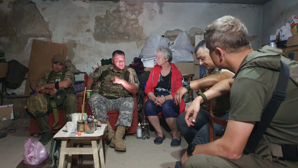 A group of people sit in a cellar in Ukraine for safety during a bomb strike. Some are in combat gear. A woman in a red cardigan who lives in the cellar sits talking to the men.