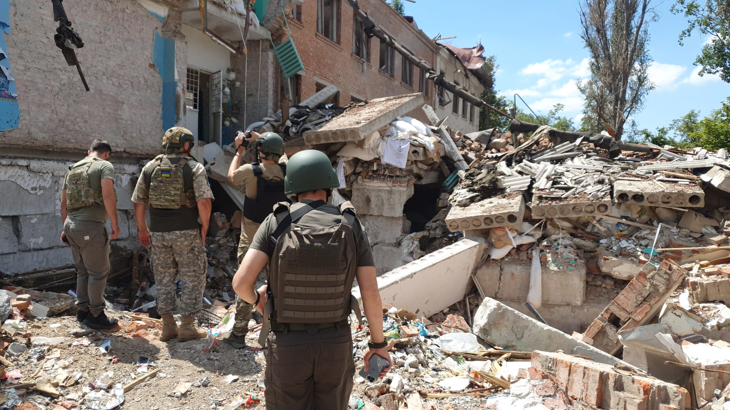 Some people in combat gear walk amongst rubble in Ukraine. The row of houses they're walking beside are half-destroyed.