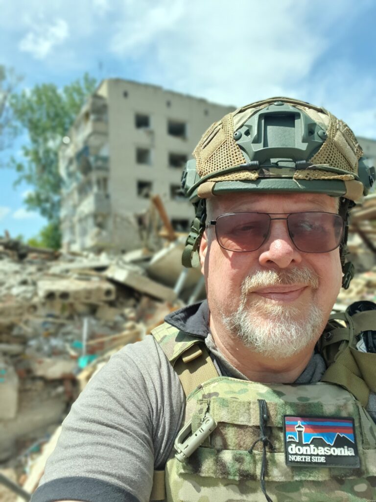 Feed The Hungry UK CEO Gwyn stands in front of a bombed building near the Ukrainian frontline. He is in full combat gear and sunglasses. Behind him is a large pile of rubble in front of a half-destroyed building.
