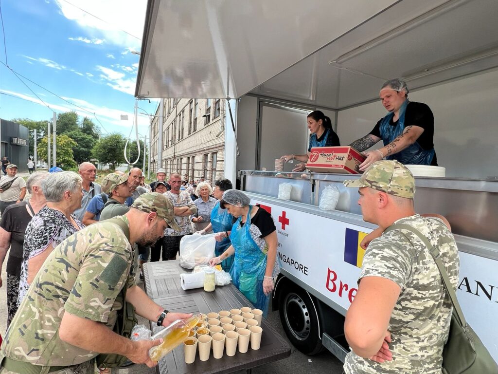 A mobile food van, set up by Feed The Hungry partner Bread of Life, serves hot food to Ukrainians living in the midst of war. The sky is blue. A queue of men and women, some in combat gear, help themselves to food.