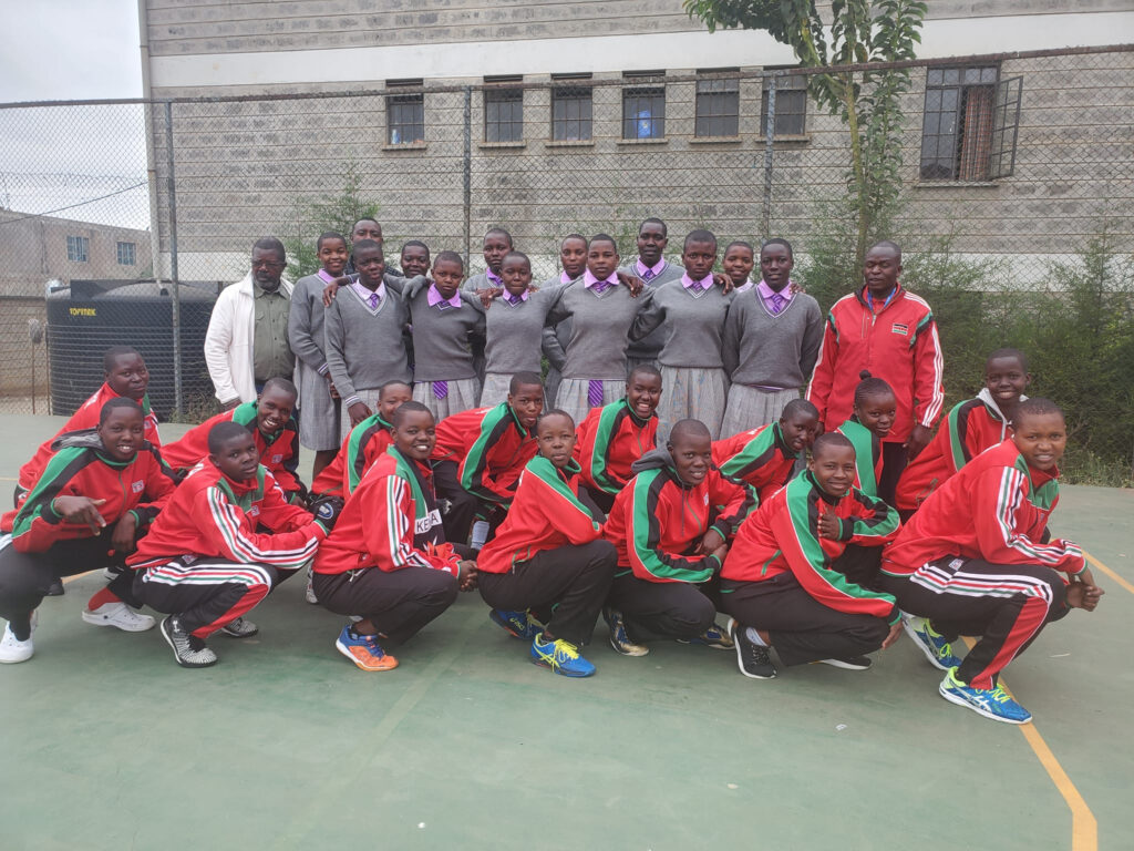 A team photo of female school age volleyball players. Some girls are in school uniform and others in red and navy sports kits. They're all smiling.