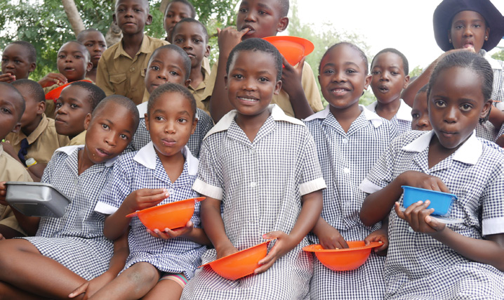 A group of school girls and boys in Zimbabwe smile at the camera while they eat nutritious rice meals from orange plastic bowls.