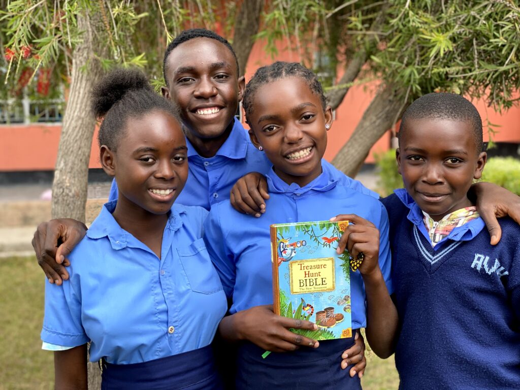 Two school girls and two school boys in Zambia smile happily at the camera. One of them is displaying a Bible they received in school. They are all wearing blue school uniform. Behind them is the red mud wall of a school building.
