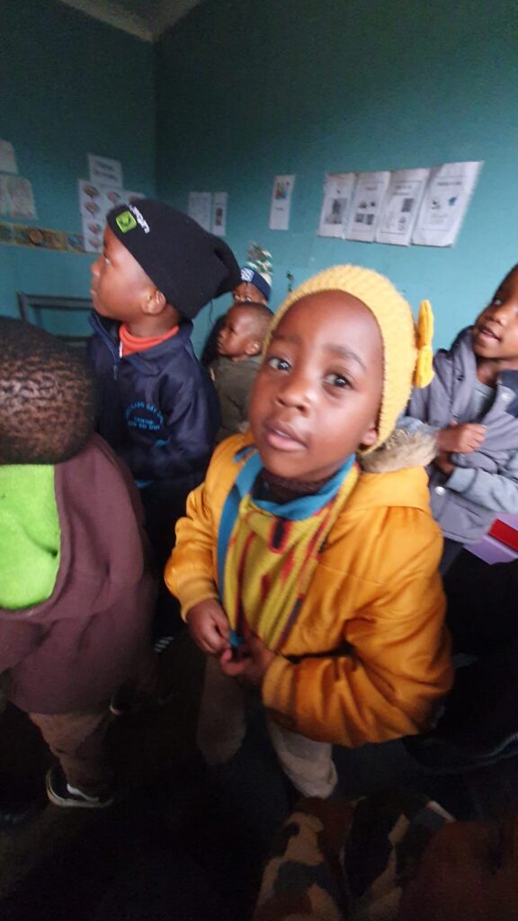 A young South African boy, wearing a yellow hat and yellow jacket, looks up at the camera. He has a half smile on his face.
