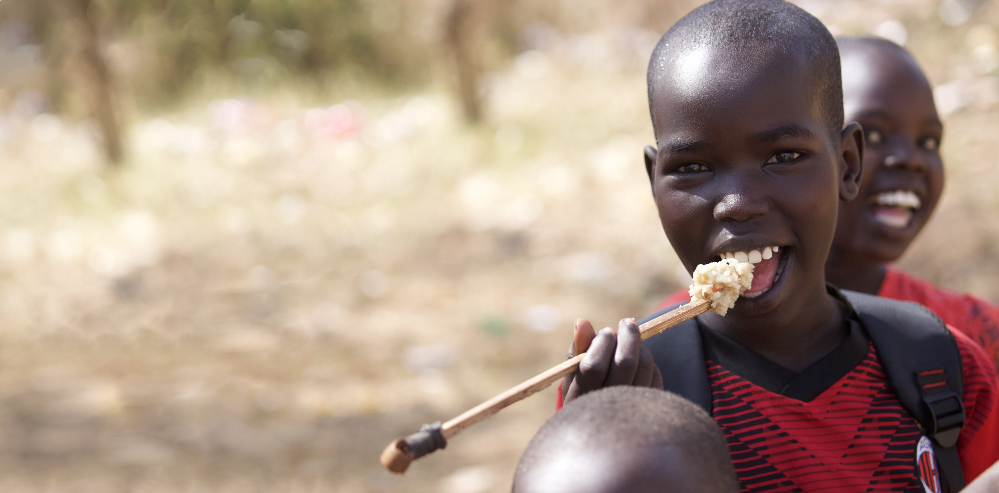 A young Ugandan boy eats rice using a stick for a spoon. In the background, another child is laughing.